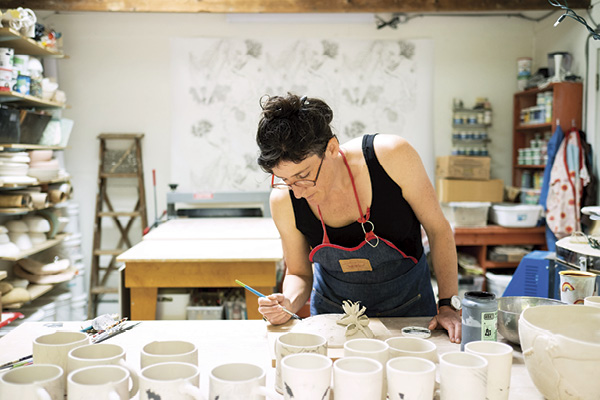 Marney McDiarmid standing at a work table, using a paintbrush to glaze a ceramic piece.