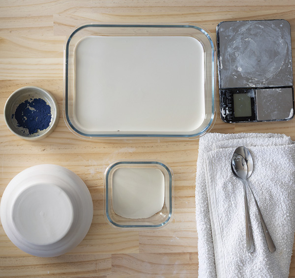 1 Gather bisqued pieces, engobe, ceramic stain, mixing bowls, a shallow tray, a teaspoon, a scale, and a towel.