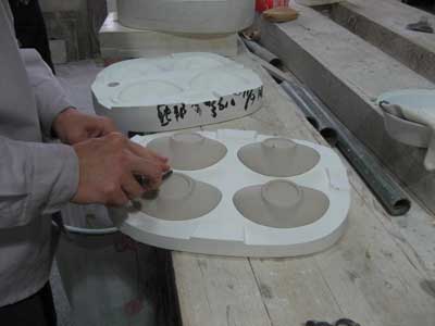 The saucers are pressure cast, a process in which very thick slip is forced into this gang mold under high pressure, and is released through vent holes on the bottoms of the saucers. Pressure casting allows for greater detail and density in the final casting. Here, the slip that is pushed through the small vent holes is being removed before de-molding.
