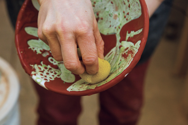 7 After the wax patterns are dry, green glaze is poured over any remaining bare clay and wax designs. Clean the glaze drips off of the waxed areas with a sponge before the bowl is fired again.