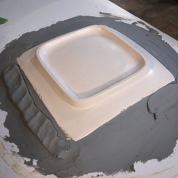 1 Part 1 of a die casting of the exterior of a square plate in progress. Clay is built up to remove undercuts before pouring Ceramical (a type of very hard, dense gypsum and concrete only used to make dies for RAM pressing).