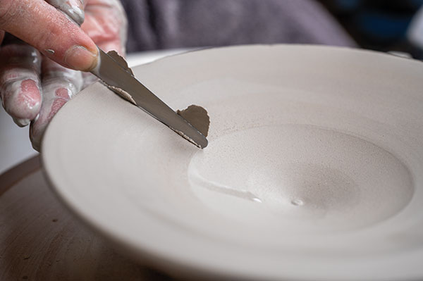 9 Throw an upside-down bowl for a lid. Lay it down with the flat side of a fettling knife, while the other hand supports the clay.