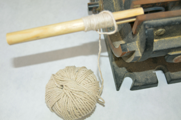 Vise holding a wood rod fitted with a metal tip while string is wrapped around and glued to hold the string in place.